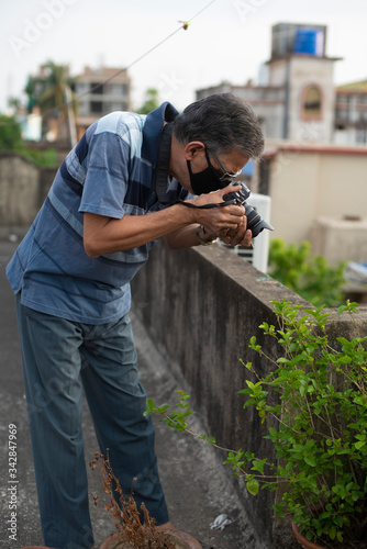 Portrait of an Indian old man wearing corona preventive mask taking photograph of a plant on a rooftop in home isolation in green background.Indian lifestyle, disease and home quarantine.