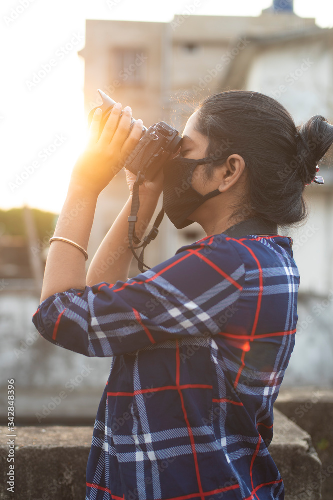 Back light portrait of an Indian young woman with corona preventive mask taking photographs on a rooftop in home isolation.Indian lifestyle, disease and home quarantine.