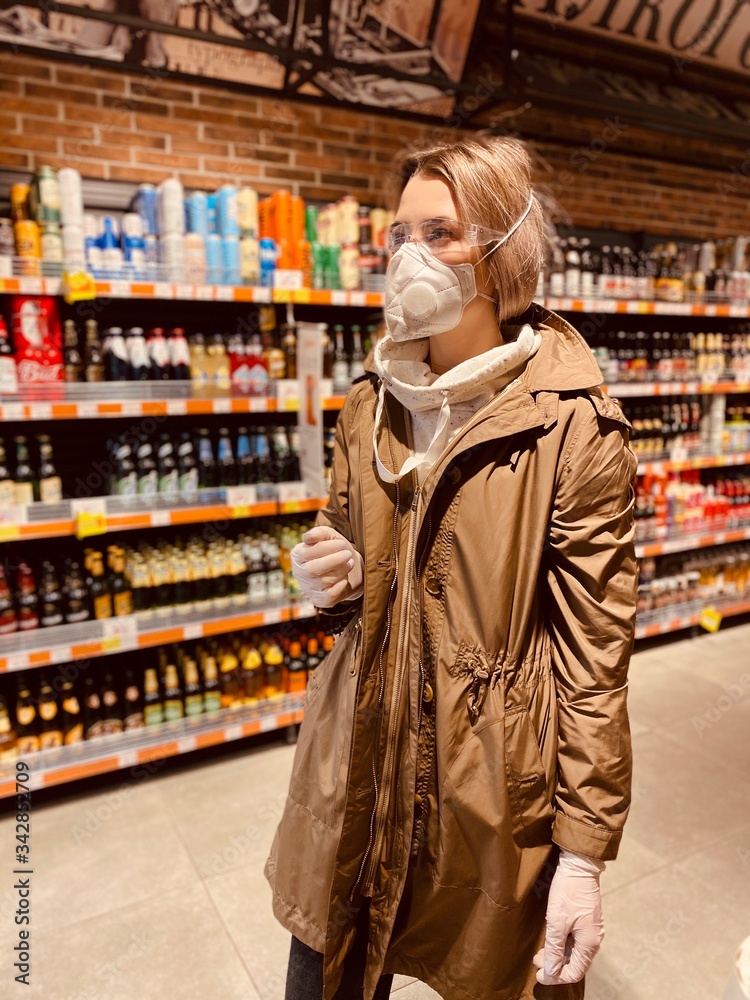 woman shopping in supermarket. beautiful girl in a protective flu mask, goggles and gloves in a store, holds goods, purchases during quarantine - against coronavirus