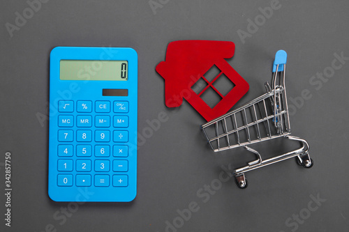 Calculation of the cost of buying or selling a home. Calculator, figurine of a house in a shopping trolley on gray background. Top view. Flat lay