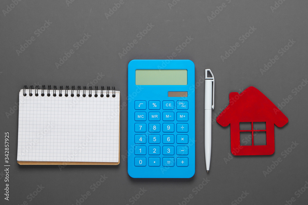 Planning and calculation of the cost of rental housing. Red house figurine, calculator, notebook on gray background. Top view Planning and calculation of the cost of rental housing. Red house figurine, calculator, notebook on gray background. Top view