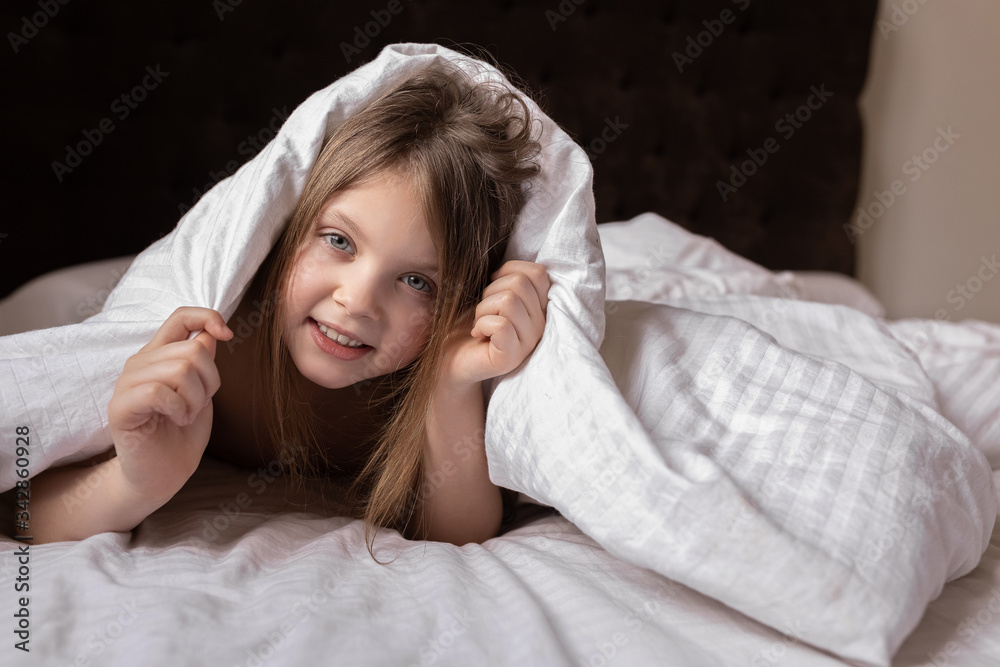 A little girl looks out from under the covers on the bed at home. Portrait of a cute smiling little blonde in a blanket, looking directly at the camera, hiding from her parents A little girl looks out from under the covers on the bed at home. Portrait of a cute smiling little blonde in a blanket, looking directly at the camera, hiding from her parents