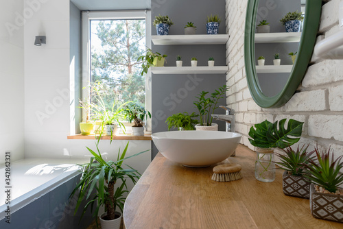 White bathroom with window and green plants. Cozy interior with wooden counter  brick wall  ceramic sink and modern design. Jungle in a bathroom.