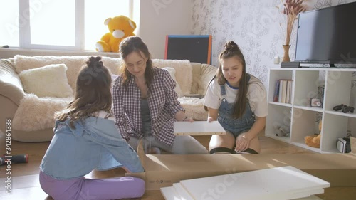 A mother and two daughters together unpack a box with a new rack for the living room.