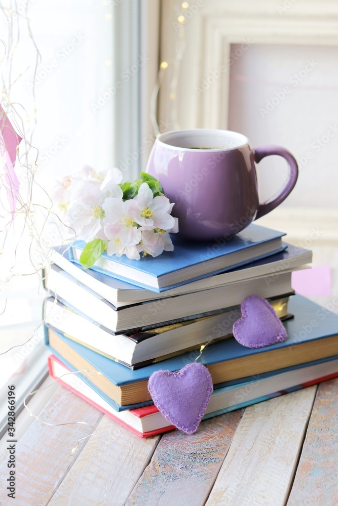 Naklejka premium Cup of coffee, a stack of books decorated with hearts, apple flowers on a wooden windowsill, the concept of home comfort
