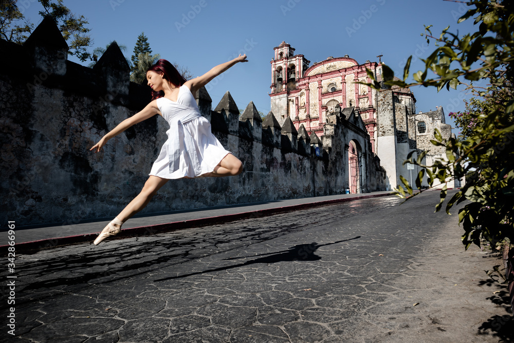 Obraz premium Young girl extend her arms and legs when dancing and jumping wearing flats ballet and white dress in old and empty street of México in front of old church under a blue sky on a spring morning.