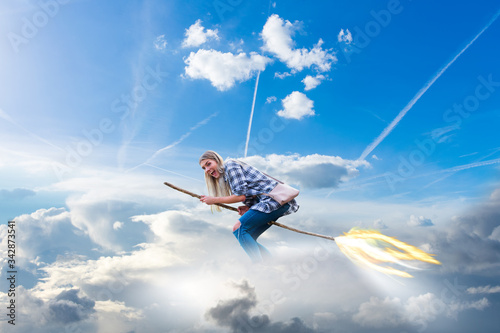 junge, modern gekleidete Frau fliegt auf einem Hexenbesen mit Feuerschweif durch die Wolken