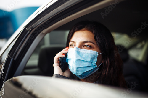 Woman wearing a medical sterile mask in a taxi car on a backseat looking out of window talking on the phone. Girl passenger waiting in a traffic jam during coronavirus quarantine. Healthcare concept.