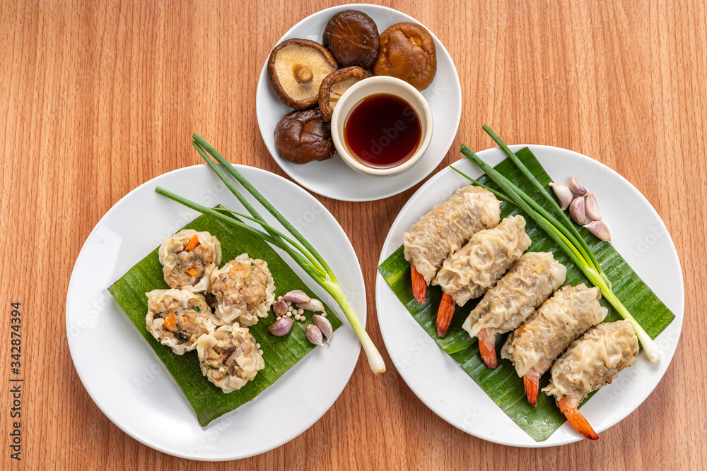 Close up Asian Steam Dumplings or Dim Sum a Famous Chinese Food with Pork and Shrimp on Banana Leaf and White Dish on Wood Table with Spciy Ingredient. Close up Asian Steam Dumplings or Dim Sum a Famous Chinese Food with Pork and Shrimp on Banana Leaf and White Dish on Wood Table with Spciy Ingredient.