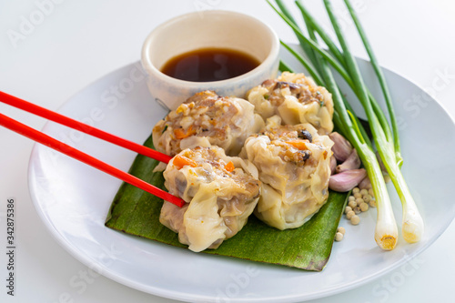 Close up Chopsticks Picking Asian Steam Dumplings or Dim Sum a Famous Chinese Food with Pork and Shrimp on Banana Leaf and White Dish with Garlic, Spring Onion and Peppers on Isolated Background.