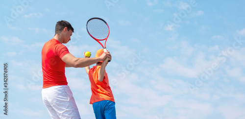 A young tennis coach or instructor teaches a child tennis player service techniques. Kids tennis on the court. Tennis school or club. Blue sky background. Banner size. Copy space for text. © Elena