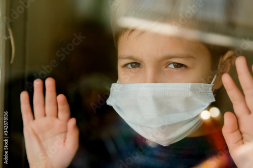 Young boy in a medical mask looks out the window. Self-isolation in quarantine, coronavirus, covid 19.