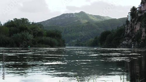 Ribera del río Ebro en la localidad de Miravet. La corriente pasa tranquilamente cruzando un paisaje de arboles y montañas en un día con alguna nube pasajera.