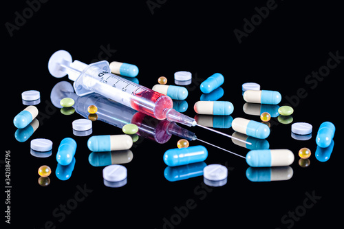 5 ml plastic medical syringe with a stainless steel needle filled with red liquid (blood) and different multi-colored tablets, pills and capsules on an isolated black background with reflection