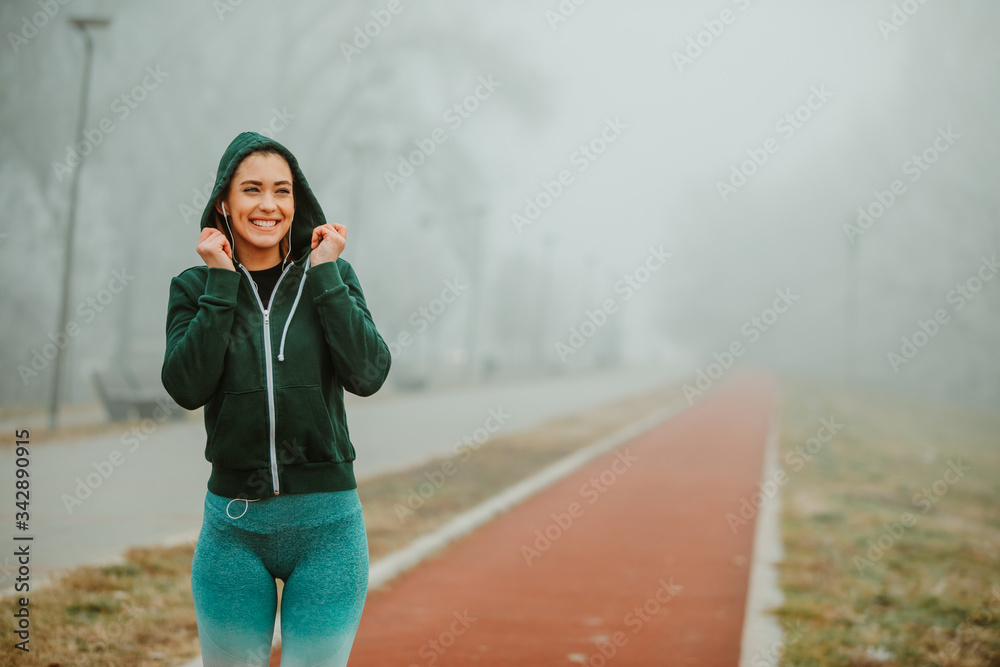 Obraz premium Fit girl in sportswear with headphones and green hoodie posing in front of camera on running track on foggy day.