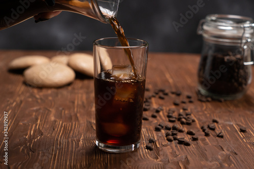 Glass of cold brew iced coffee on brown wood table with coffee beans and cookies on black background