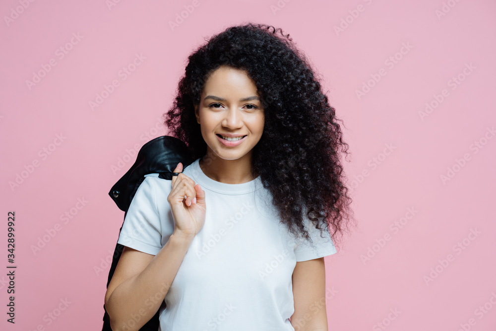 Portrait of positive ethnic woman wears white t shirt, carries jacket on shoulder, has cheerful expression, ready for picnic, enjoys day off, poses indoor against rosy background. People, lifestyle