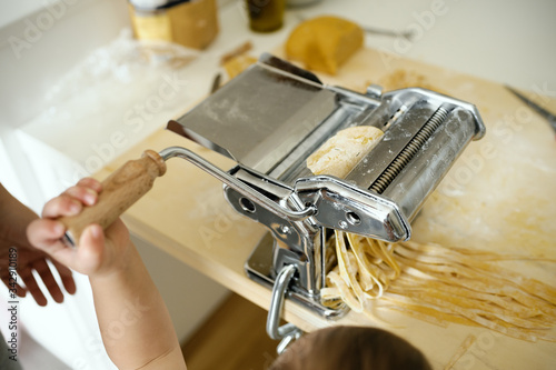 Staying at home with your family and preparing fresh home-made pasta (tagliatelle): mom cutting sheets of pasta with the help of her young son, with pasta machine on a wooden board.