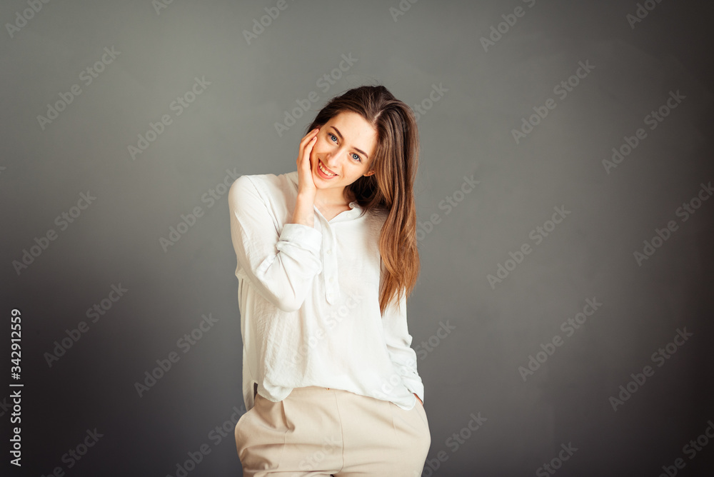 Young smiling girl in a white shirt, head tilted to the side, hand near the cheek, the second in the pocket, insulator on a gray background. No retouching. Without makeup. Young smiling girl in a white shirt, head tilted to the side, hand near the cheek, the second in the pocket, insulator on a gray background. No retouching. Without makeup.