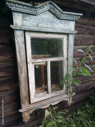  old carved window in a wooden house overgrown with nettles