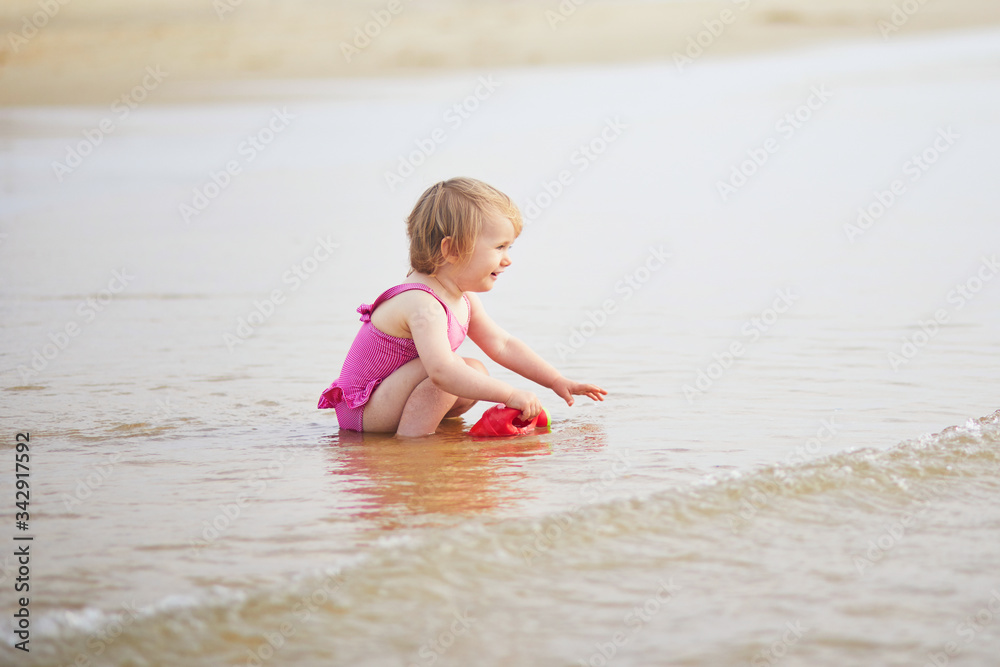 Adorable toddler girl playing with red watering can on the ocean beach Adorable toddler girl playing with red watering can on the ocean beach