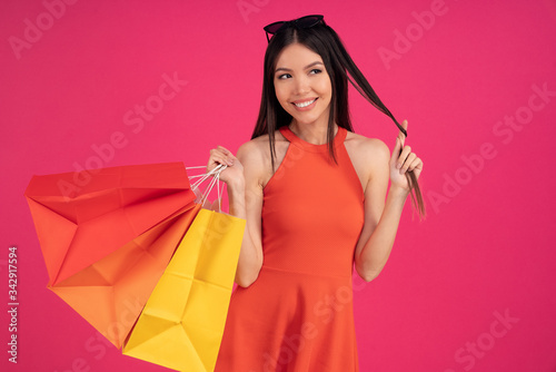 Image of a beautiful excited happy asian woman posing isolated over pink wall background holding shopping bags. dressed in orange dress and sunglasses