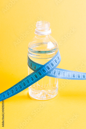 blue measure tape wrapped around a small bottle of water on a yellow background copy space, weight loss and a healthy lifestyle