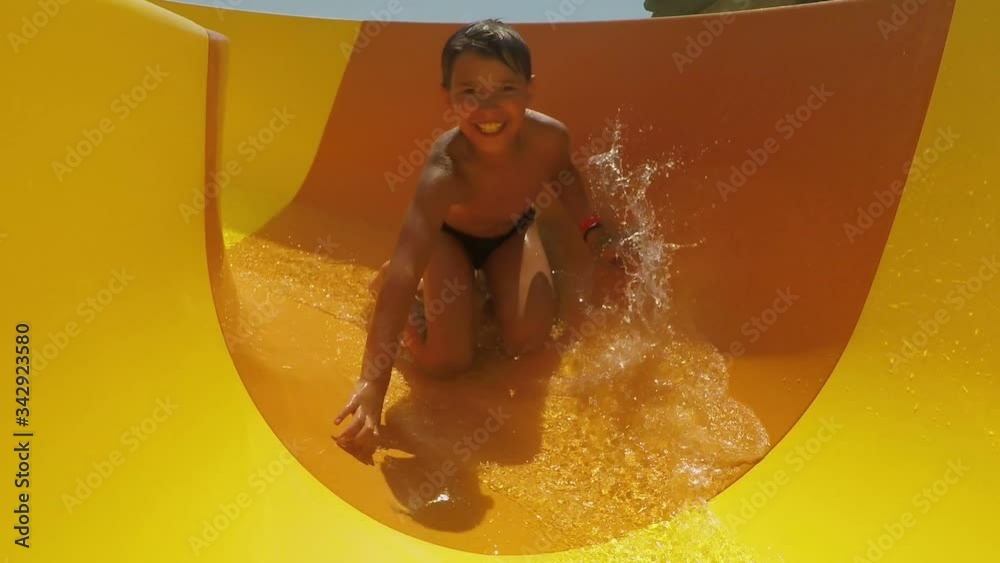 Laughing boy rides down orange slide in a water park. Boy in a swimsuit laughs and slides into the water raising spray