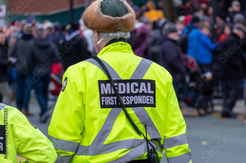 A male medical first responder walks along a street alone with people standing in the distance. The officer is wearing a bright yellow coat with grey reflective bands, black pants and a radio.