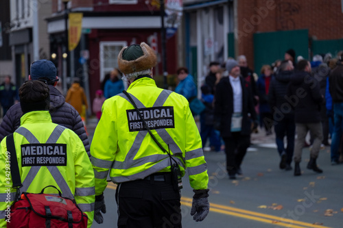 Two emergency first responders walking along a street with people and buildings in the background. The female EMT has a red first aid bag hanging off her shoulder. The tall male is carrying a radio. 
