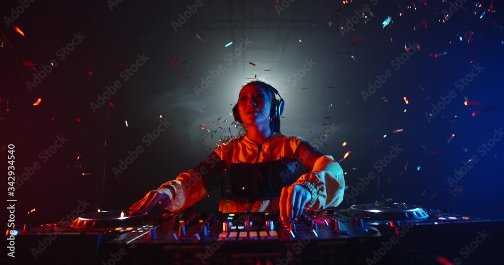 Cool asian female disc jockey standing at mixer cotroller, creating a music list in a nightclub, lit by red and blue lights - nightlife concept 4k footage