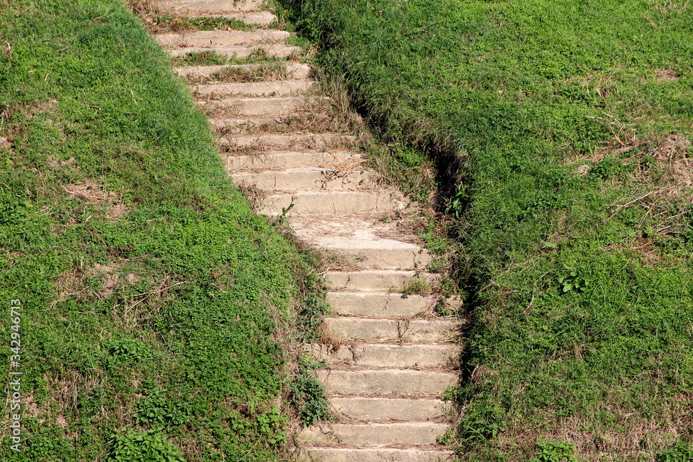 Wavy old partially broken cracked concrete steps surrounded with uncut green grass mixed with dry grass on side of local river bank Wavy old partially broken cracked concrete steps surrounded with uncut green grass mixed with dry grass on side of local river bank