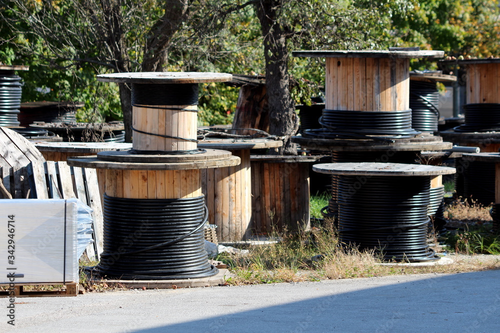 Fototapeta premium Thick black electrical cable on various sizes industrial wooden cable winding spool drum reels left at abandoned industrial complex backyard surrounded with trees and paved road