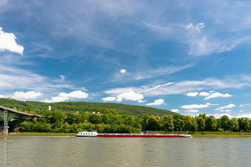 A barge flowing on the river Rhine in western Germany with a blue sky with clouds and a forest in the background.