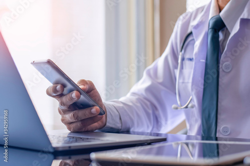 Foto Male doctor in white lab coat, hand holding and using mobile smart phone with laptop computer and digital tablet on the desk