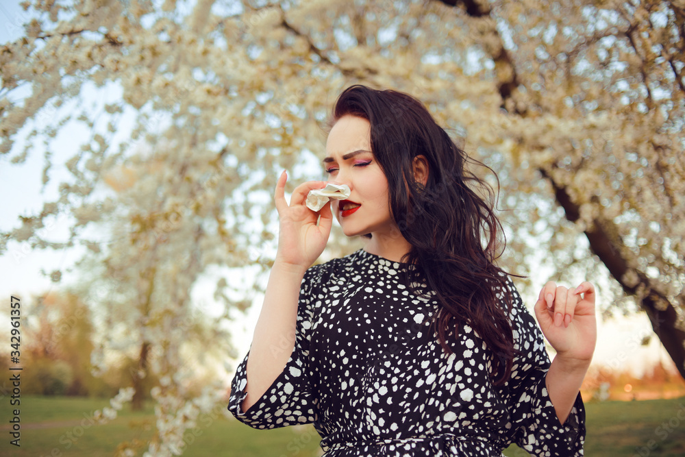 The girl blows her nose, the girl is allergic to flowering, spring. Girl sneezes into a napkin, culture, runny nose, spring, flowers. Model on a background of a tree with flowers sneezes. The girl blows her nose, the girl is allergic to flowering, spring. Girl sneezes into a napkin, culture, runny nose, spring, flowers. Model on a background of a tree with flowers sneezes.