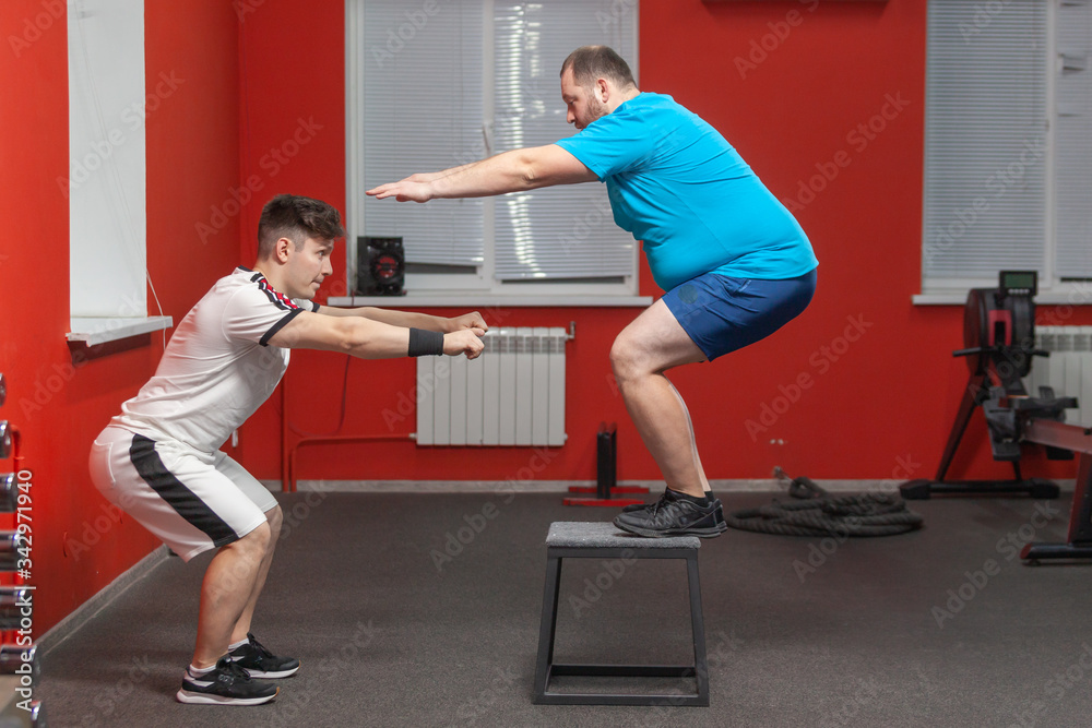 Eager overweight man is doing jumping in the gym under the guidance of