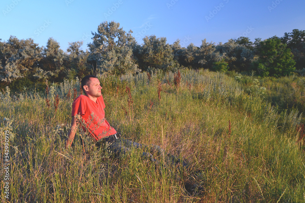 tired lonely young man in a red t-shirt and jeans relaxig in grass in the meadow near the trees at sunset	