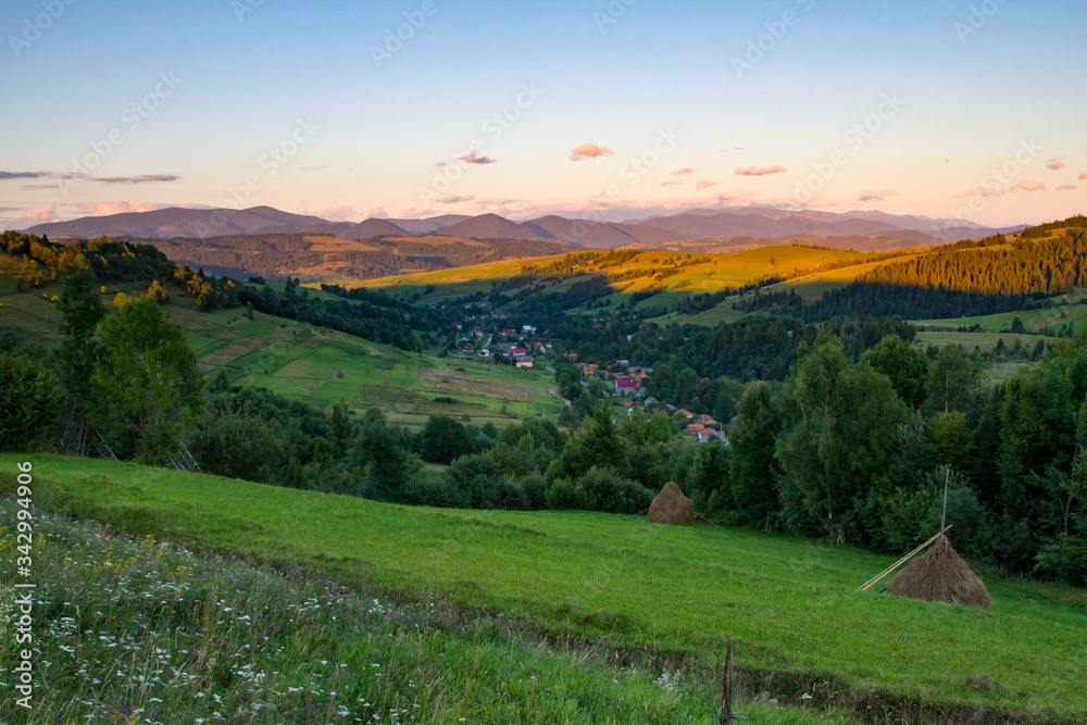Village in the mountains at sunset, mountain range in the background. Alpine meadow and haystack on it in the foreground. Carpathians