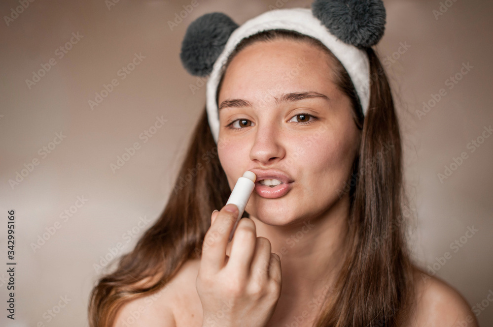 Obraz premium a brunette girl without makeup with problematic skin with a bandage with ears on her head naked wrapped in a towel after a bath doing Spa treatments applies lip balm to lips moisturizes
