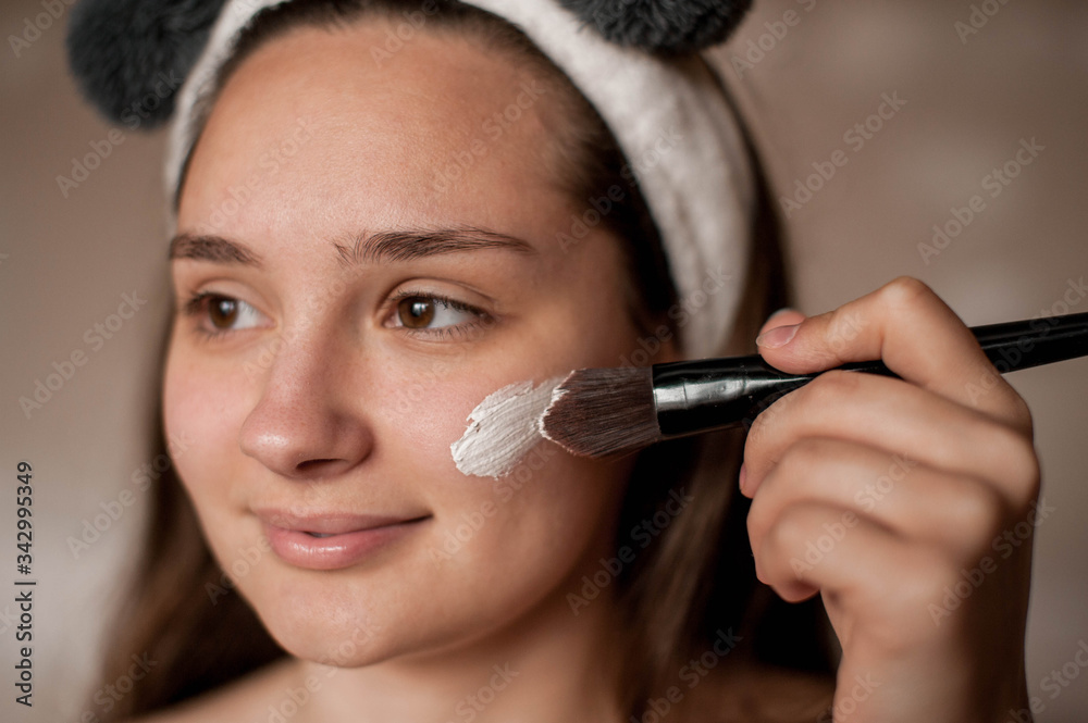 a brunette girl without makeup with problematic skin with a bandage with ears on her head naked wrapped in a towel after a bath doing Spa treatments applies a white clay mask to the face with brush a brunette girl without makeup with problematic skin with a bandage with ears on her head naked wrapped in a towel after a bath doing Spa treatments applies a white clay mask to the face with brush