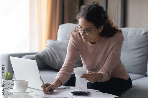 Focused millennial woman in eyewear writes out information from invoices, calculating monthly expenses alone at home. Young lady managing incomes and outcomes, planning investments or summer vacation. photo