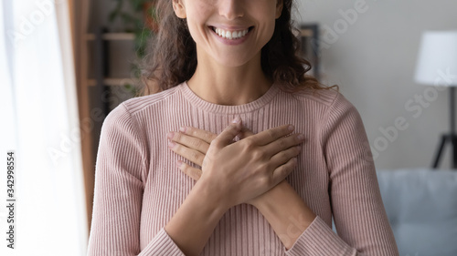 Close up focus on happy sincere female holding folded hands on chest. Emotional positive kind candid millennial woman feeling thankful indoors, showing gratitude sign, believe faith charity concept. photo