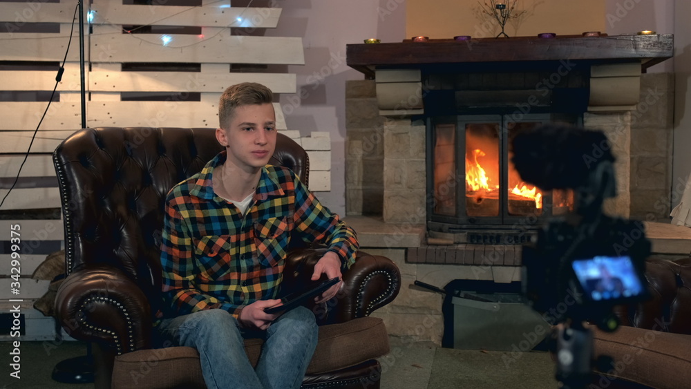 Home video shooting: teenage boy speaking something to camera with tablet in his hands. He sitting in comfortable leather armchair near the fireplace Home video shooting: teenage boy speaking something to camera with tablet in his hands. He sitting in comfortable leather armchair near the fireplace