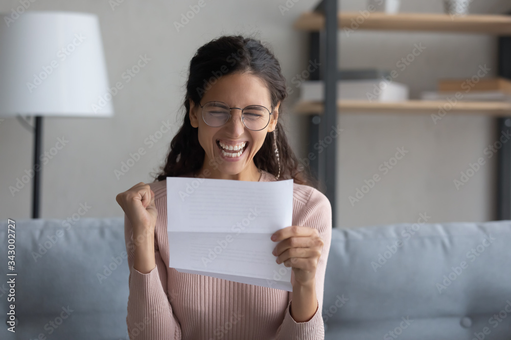 Overjoyed young attractive lady in eyewear holding paper letter, celebrating business success. Emotional millennial woman making yes gesture, feeling excited about personal achievement at home. Overjoyed young attractive lady in eyewear holding paper letter, celebrating business success. Emotional millennial woman making yes gesture, feeling excited about personal achievement at home.