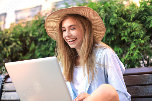 Attractive young woman wearing summer clothes working on laptop while sitting on bench in city street.