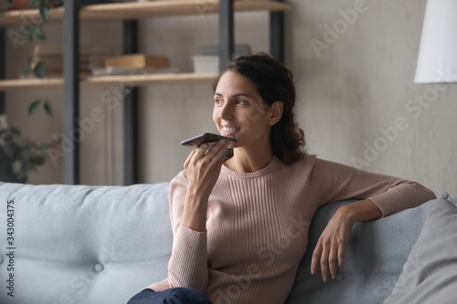 Happy young woman relaxing on comfortable sofa, sending audio message, sharing news with friends. Pretty smiling lady enjoying pleasant free weekend time alone at home, activating voice assistant.