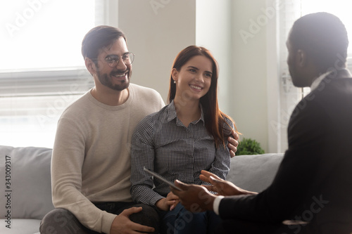 Young happy couple, man and woman negotiating purchase deal with manager. Smiling african american businessman talking with customers at meeting. Diverse seller and client in office.