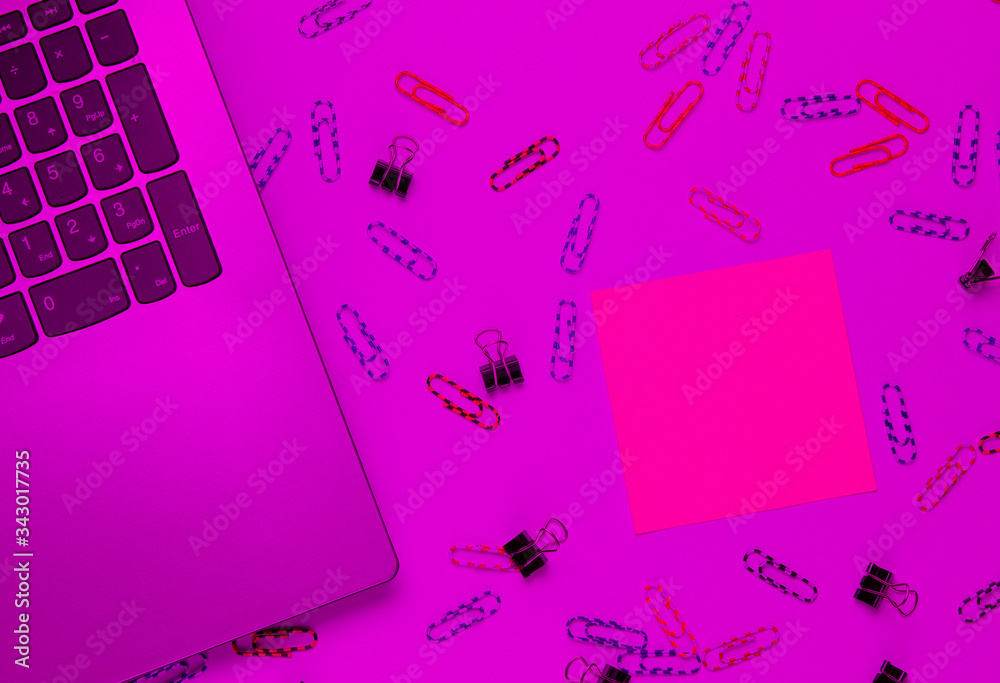 Minimalistic office still life. Laptop, stationery (paper clips, felt-tip pen, memo piece of paper) in pink neon light. Studio shot.