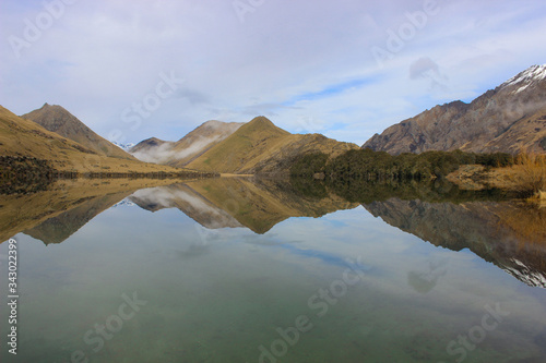 Reflections of Moke Lake, Otago, New Zealand. Still water, backed by snow capped mountains and grassy plains. 
